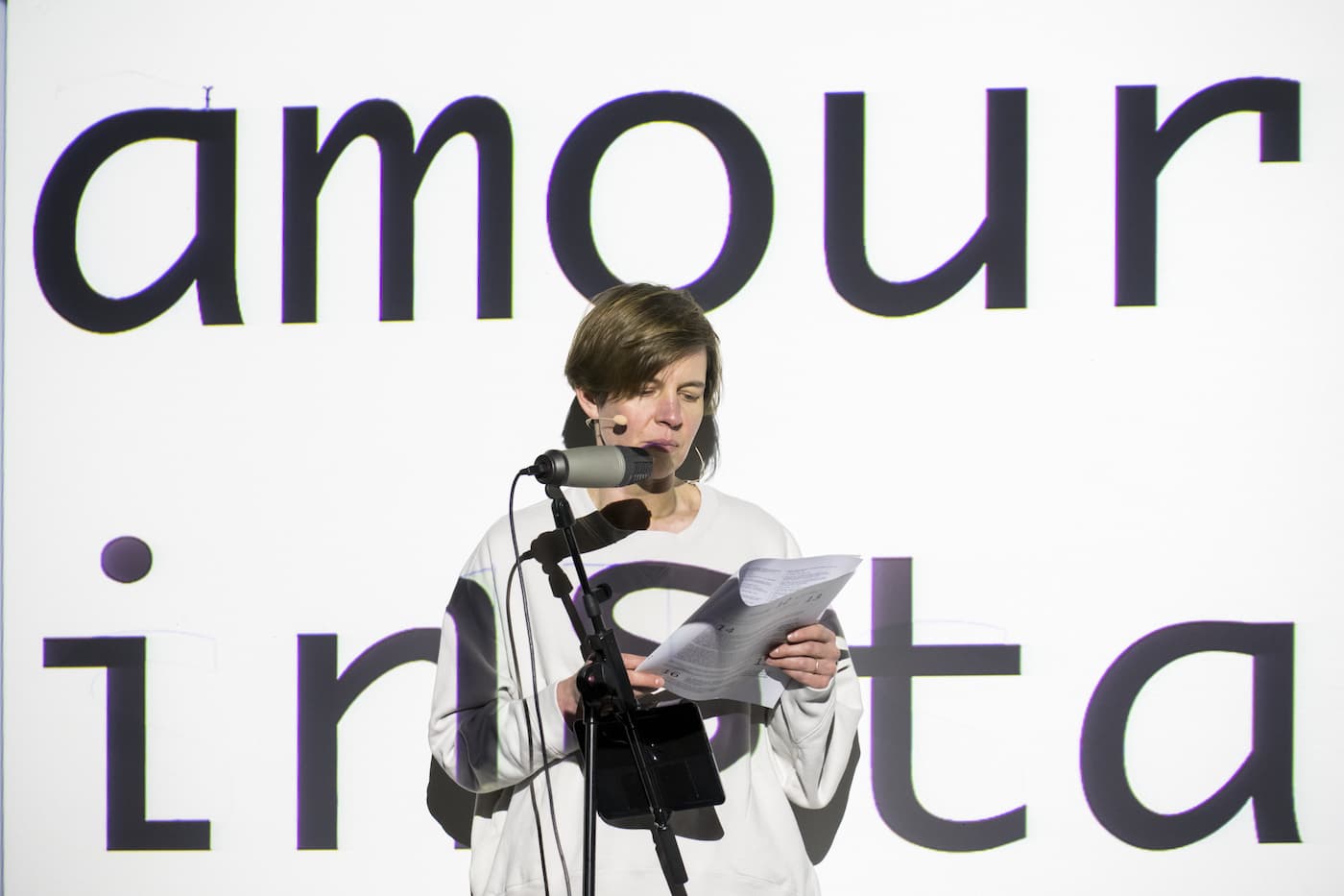 The artist Anna Barham giving a reading of one of her perfomances standing behind a microphone. A series of words are projected on top of her.