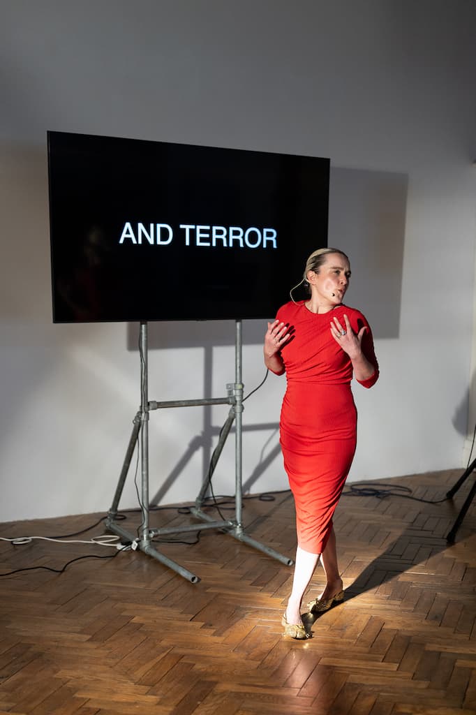 A female performer in a red dress stading in front of a screen with "AND TERROR" in large capital letters on it.