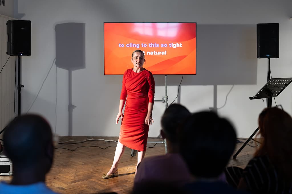 A female performer in a red dress stading in front of a screen with "to cling to this so tight and natural" in large capital letters on it.