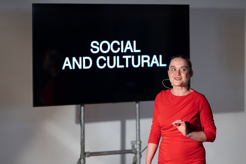 A female performer in a red dress stading in front of a screen with "SCOIAL AND CULTURAL" in large capital letters on it.