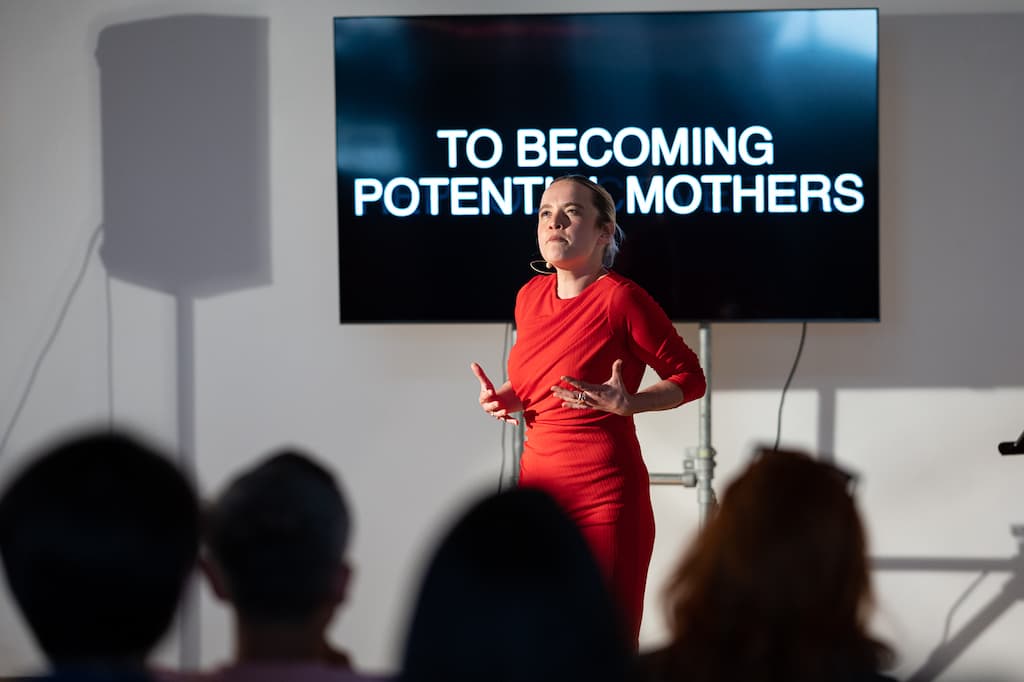 A female performer in a red dress stading in front of a screen with "TO BECOMING POTENTIAL MOTHERS" in large capital letters on it.