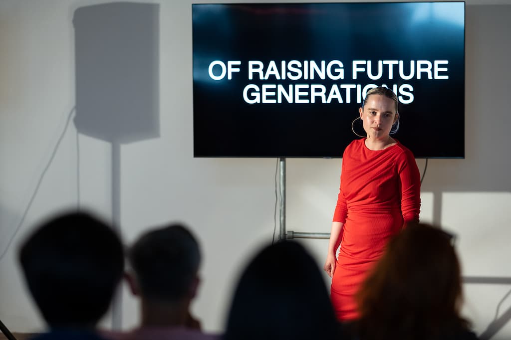 A female performer in a red dress stading in front of a screen with "OF RAISING FUTURE GENERATIONS" in large capital letters on it.