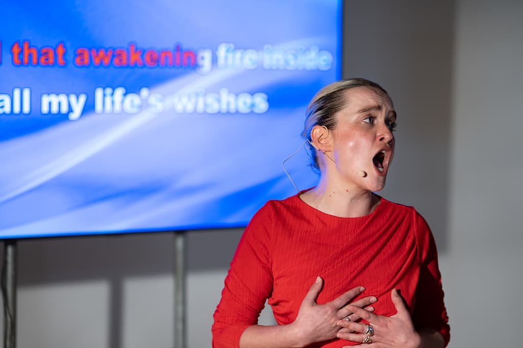 A female performer in a red dress stading (possibly singing) in front of a screen with some blurred text on it.