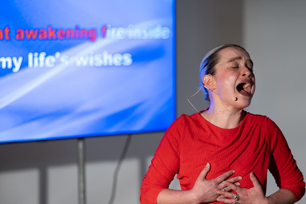 A female performer in a red dress stading (possibly singing) in front of a screen with some blurred text on it.