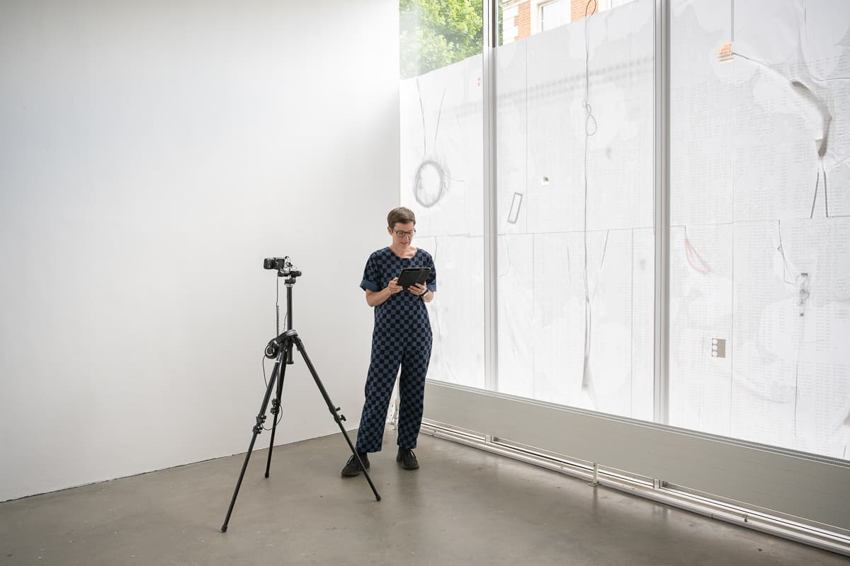 The artist Anna Barham giving a reading of one of her perfomances standing behind a microphone. A series of words are projected on top of her.