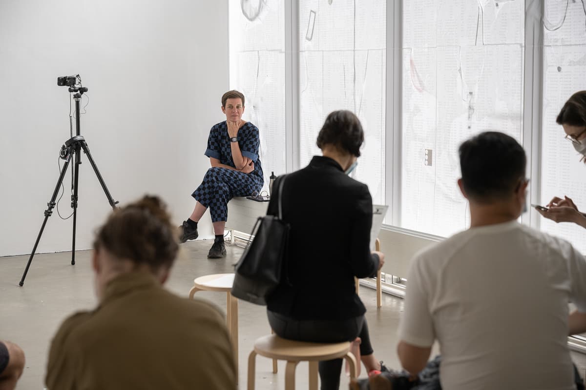 The artist Anna Barham giving a reading of one of her perfomances standing behind a microphone. A series of words are projected on top of her.