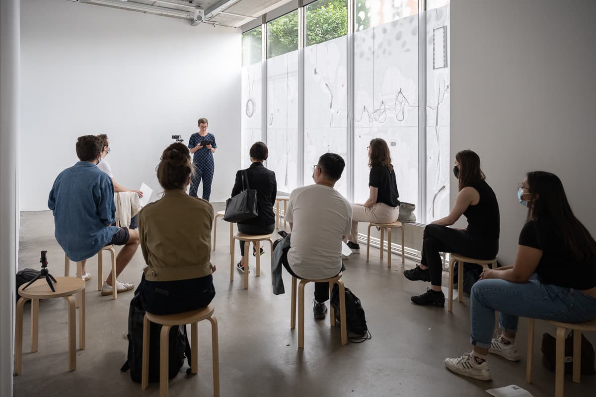 The artist Anna Barham giving a reading of one of her perfomances standing behind a microphone. A series of words are projected on top of her.