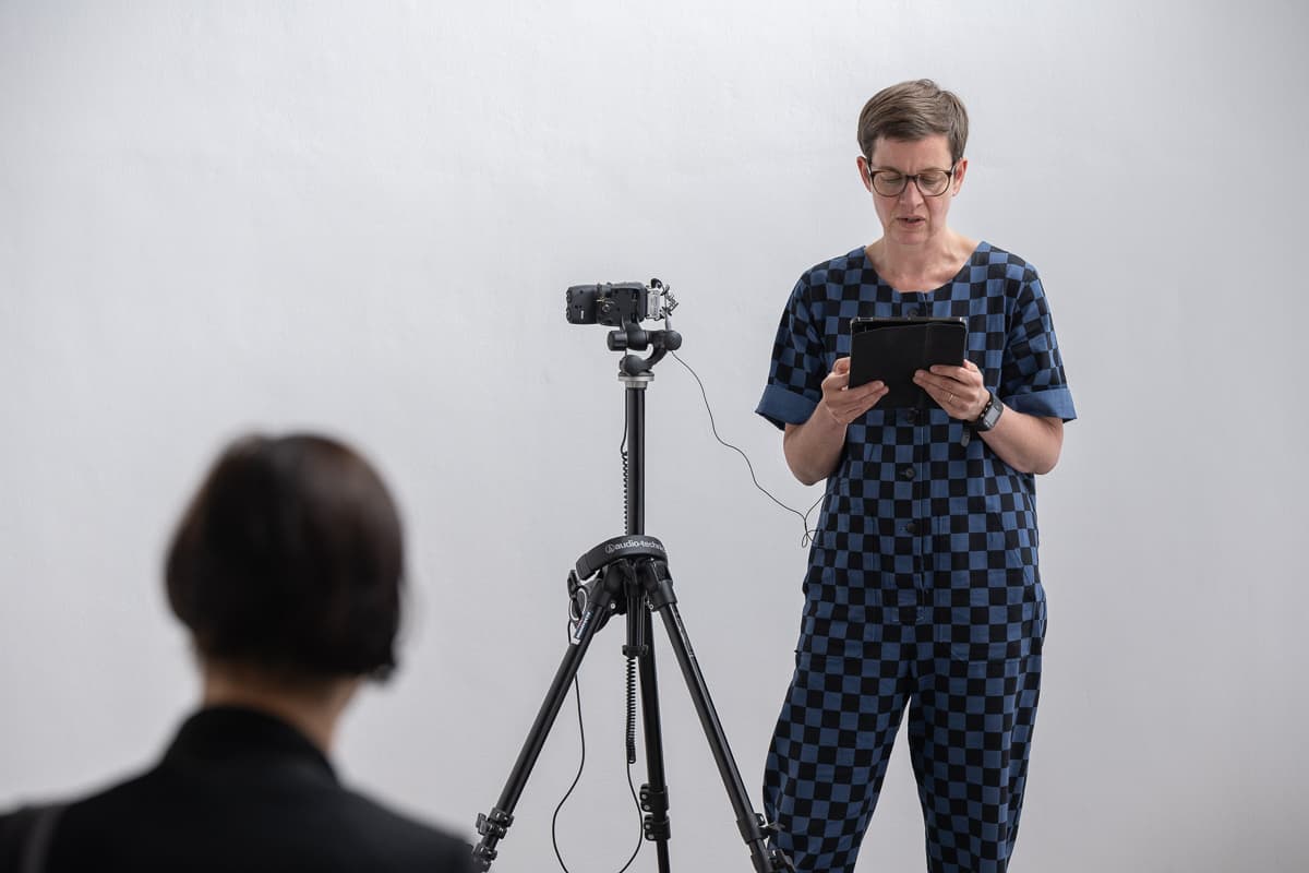 The artist Anna Barham giving a reading of one of her perfomances standing behind a microphone. A series of words are projected on top of her.