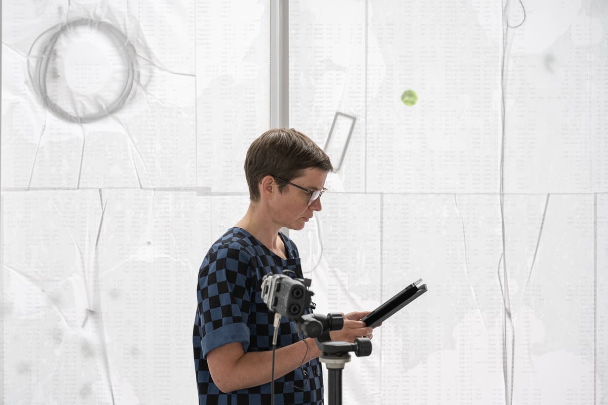 The artist Anna Barham giving a reading of one of her perfomances standing behind a microphone. A series of words are projected on top of her.