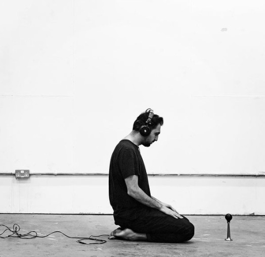 The artist Abbas Zahedi kneeling on the floor of a gallery space with headphones on.