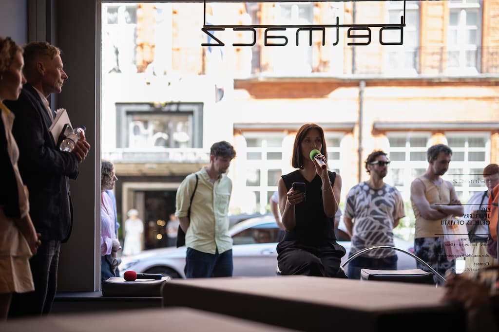 A performer sat in the window of a gallery, speaking into a green microphone, while reading a script from a phone.