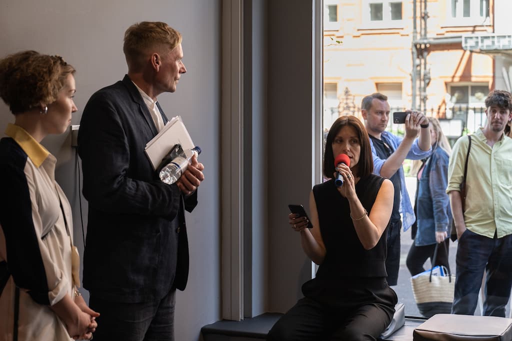 A performer sat in the window of a gallery, speaking into a red microphone, while reading a script from a phone.