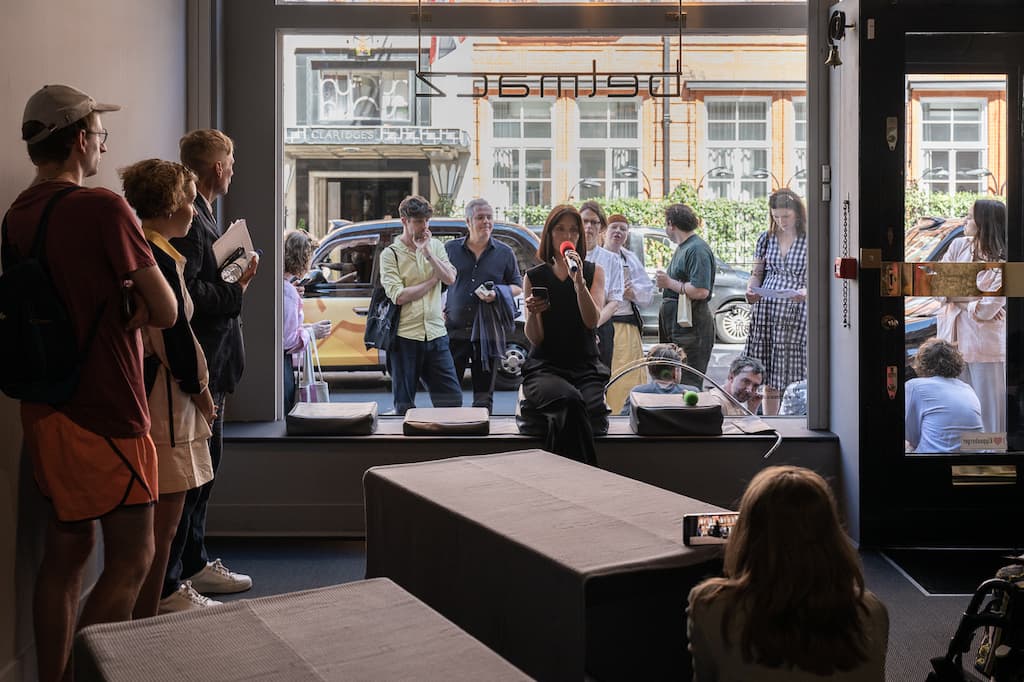 A performer sat in the window of a gallery, speaking into a red microphone, while reading a script from a phone.