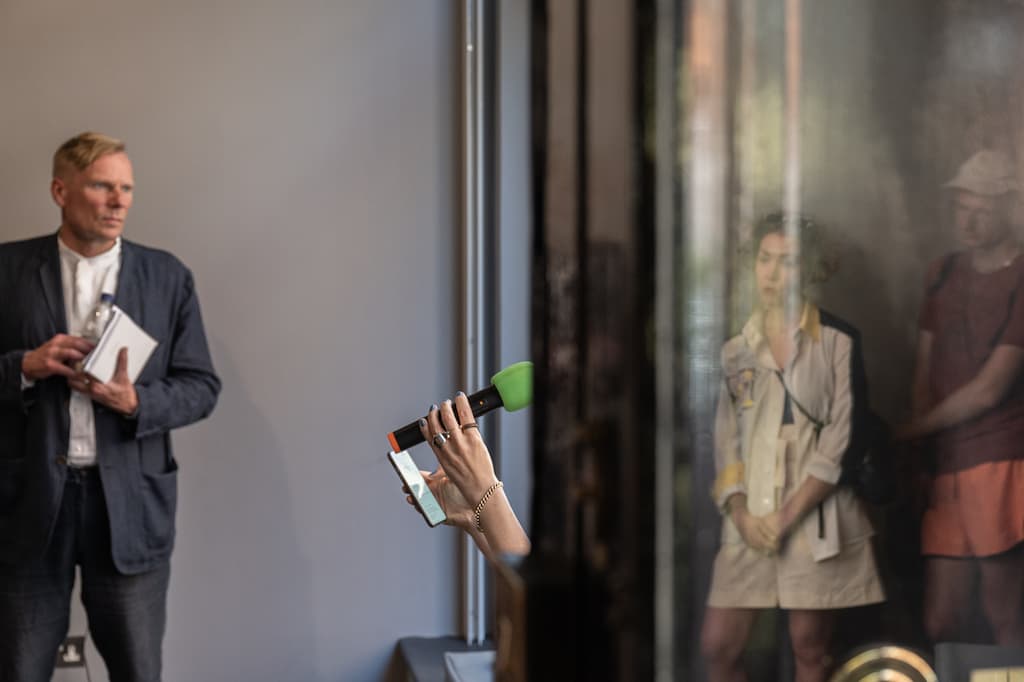 Side biew of a performer sat in the window of a gallery, speaking into a green microphone, while reading a script from a phone.