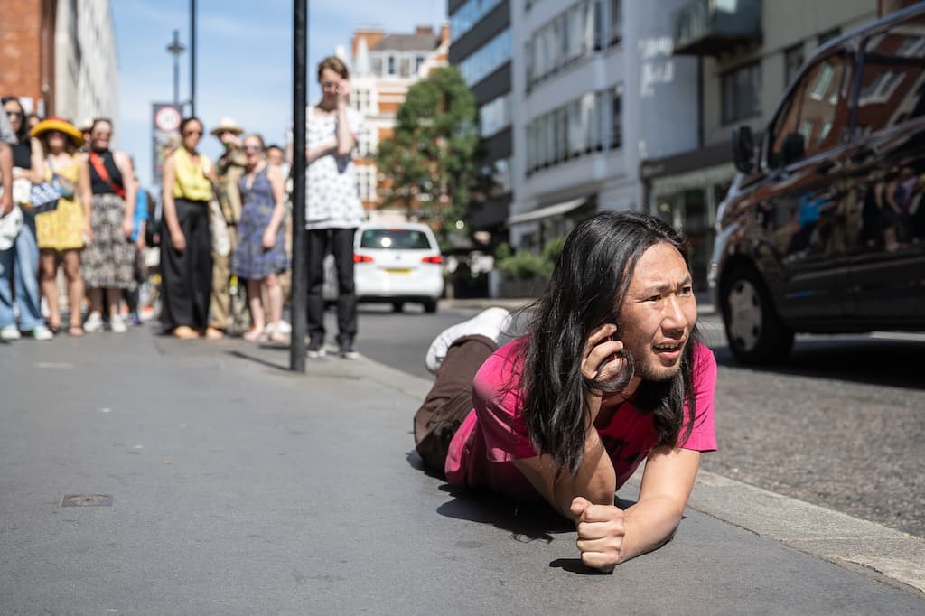 A performer laying on their front on a pavement outside a gallery while on the phone