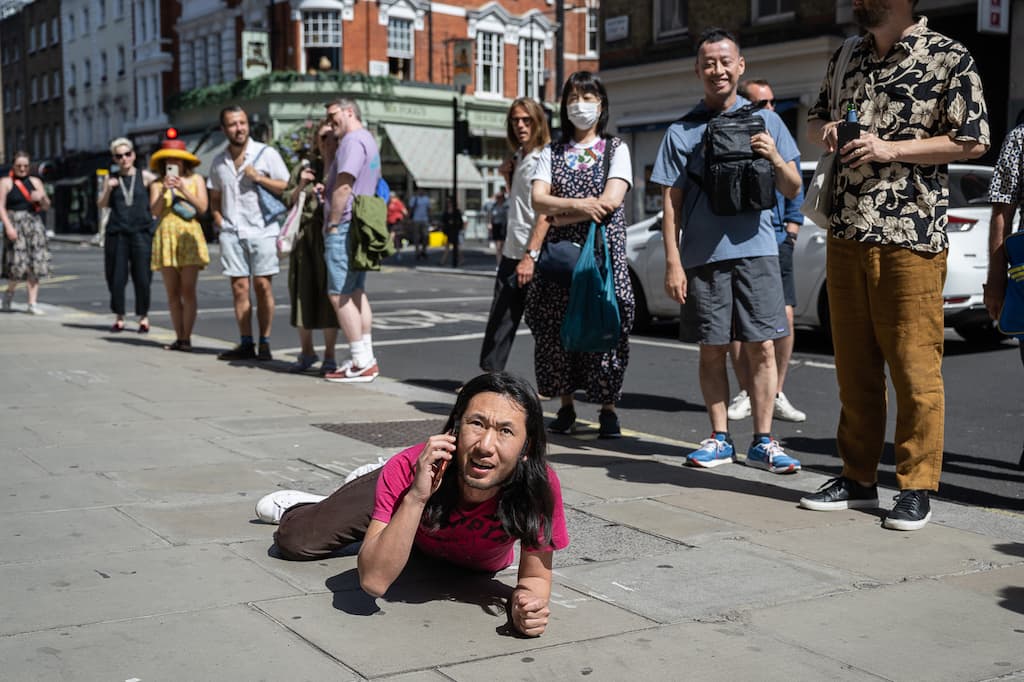 A performer laying on their front on a pavement outside a gallery while on the phone - with audience in the background.