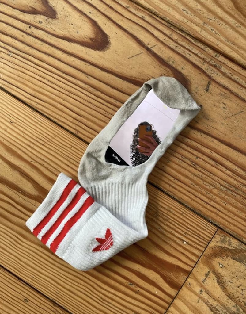 An white Addidas sock on a wooden floor with a hole in it. In the hole there is a mirror, in which you can see someone taking a picture of it with their phone