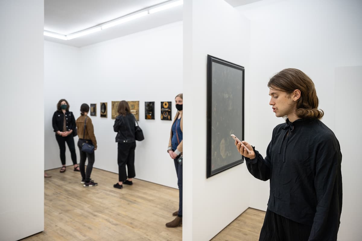 The artist Heman Chong standing in a gallery space reading from a smart phone - performing Everything (Wikipedia)