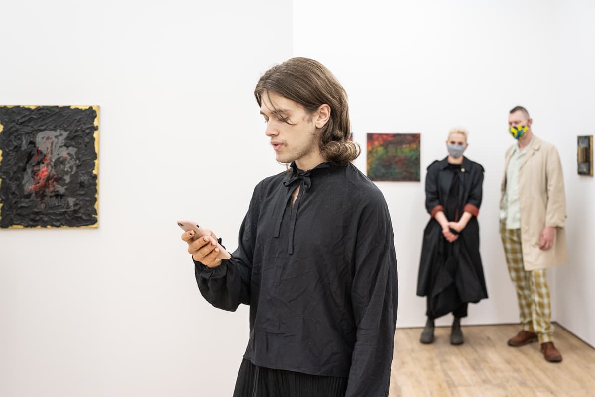 The artist Heman Chong standing in a gallery space reading from a smart phone - performing Everything (Wikipedia)