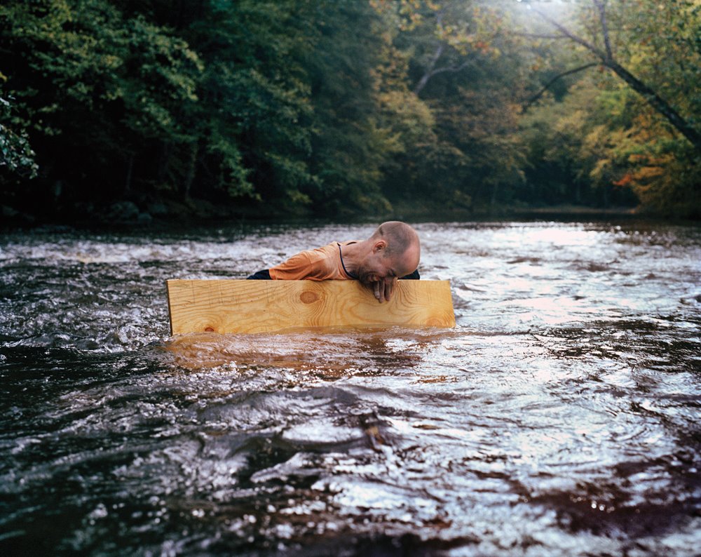 The artist Joel Kyack in a river, holding on to a piece of wood to keep afloat.