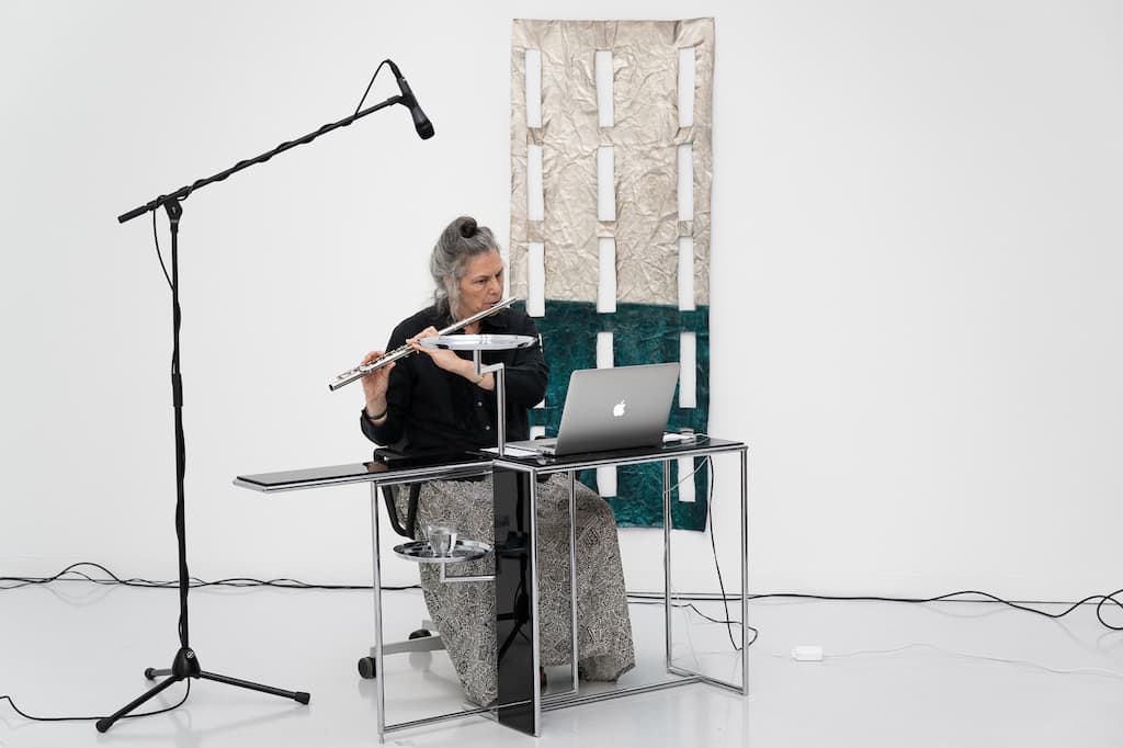 Performer playing the flute while sat behind a desk with laptop and microphone.