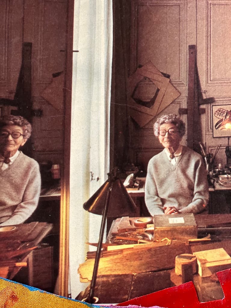 Portrait of Eileen Gray sat in her studio (or workshop). Her reflection in the mirror takes up the left side of the image.