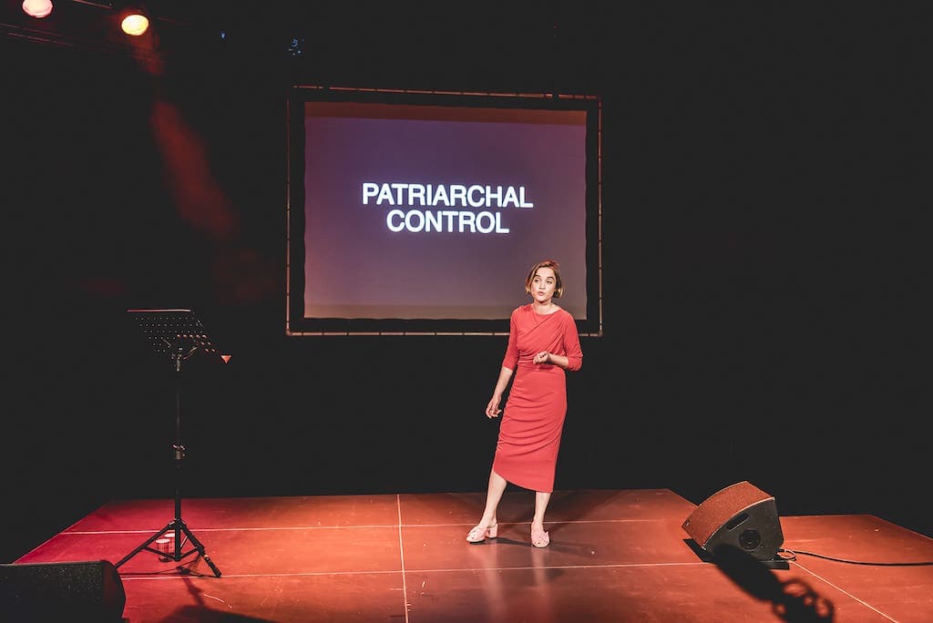 A female performer in a red dress stading in front of a screen with Patriarcal Control in large capital letters on it.