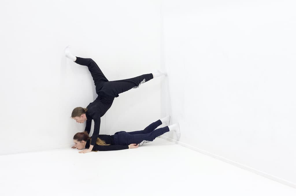 2 performers, dressed in black, one on the floor, the other doing a handstand on top, enacting A Cricle Whispering Dot by Nicole Bachmann