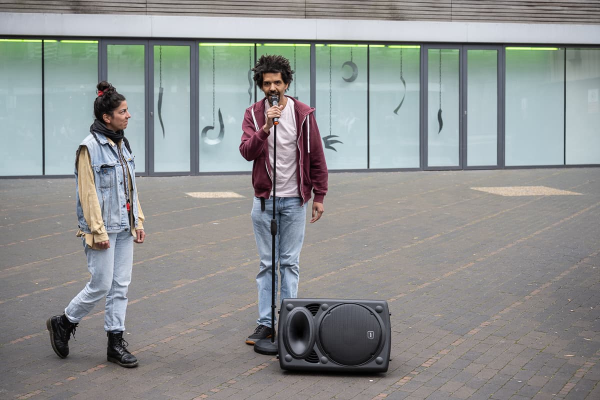 A performer with a microphone and a sub-woofer standing outside VITRINE Gallery.