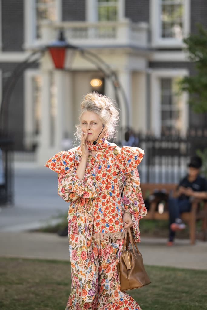 A performer in a floral dress smoking a cigarette while standing in a park.