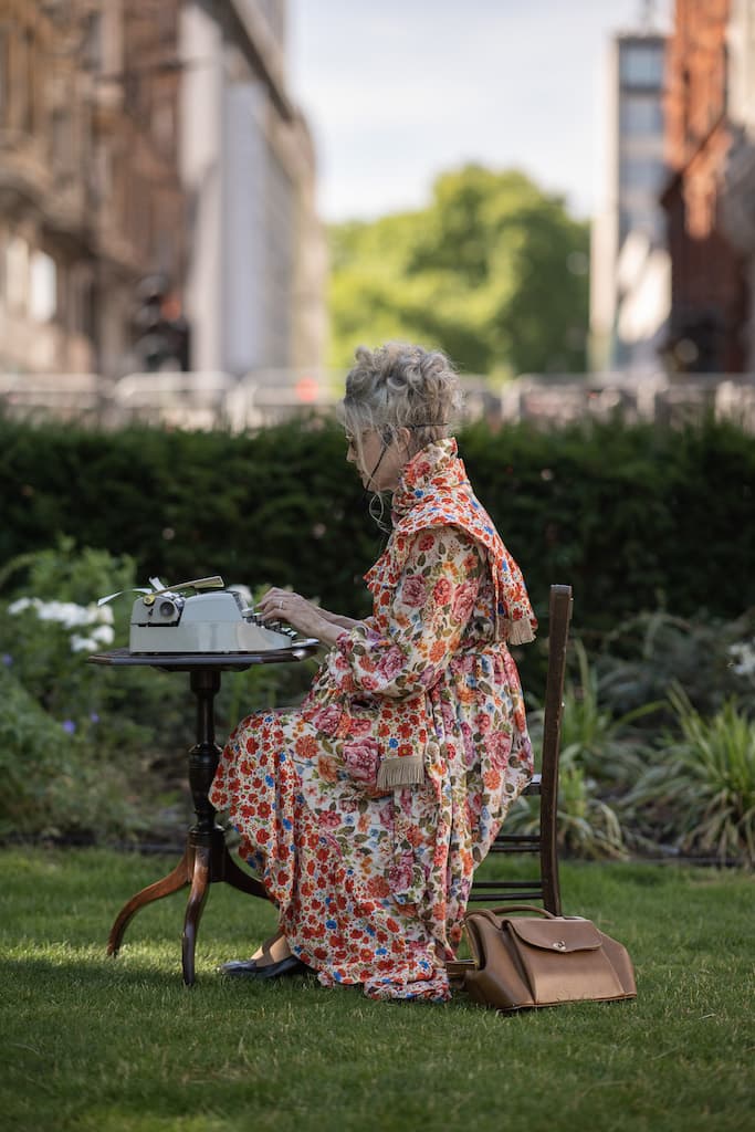 A performer in a floral dress sat at a wooden chair and desk in the middle of a park, typing on a typewriter.