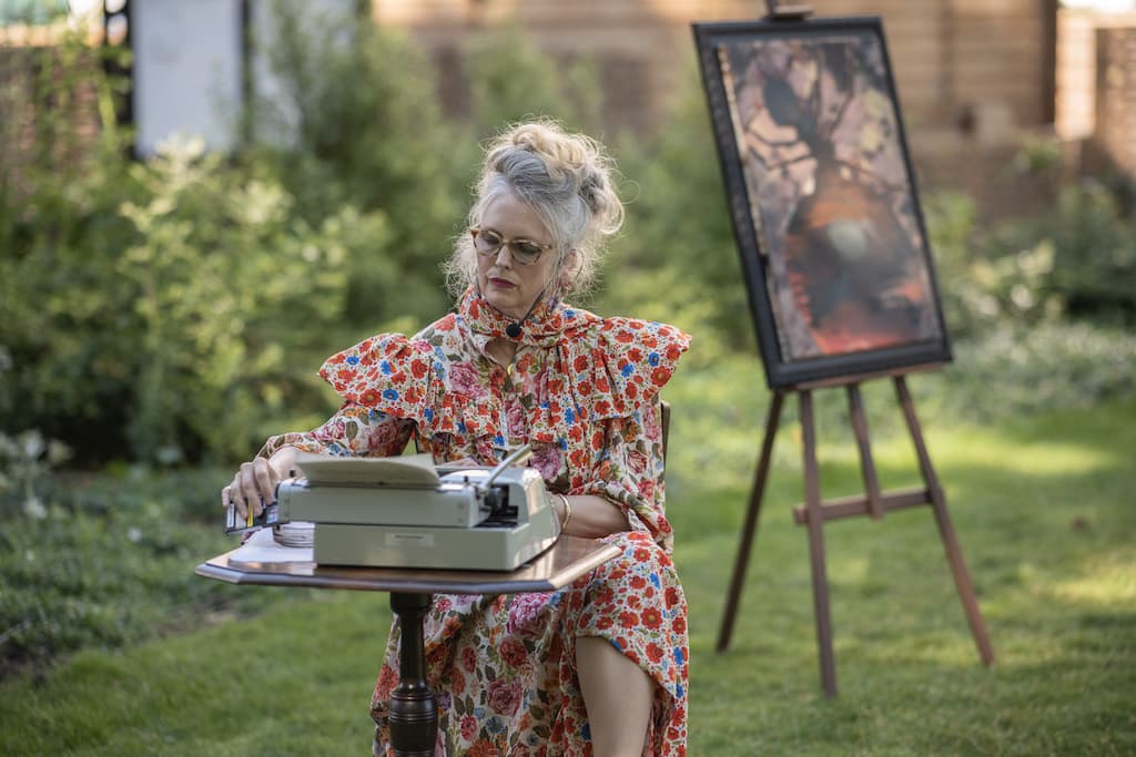 A performer in a floral dress sat at a wooden chair and desk in the middle of a park, typing on a typewriter with a painting on an easel in the background.