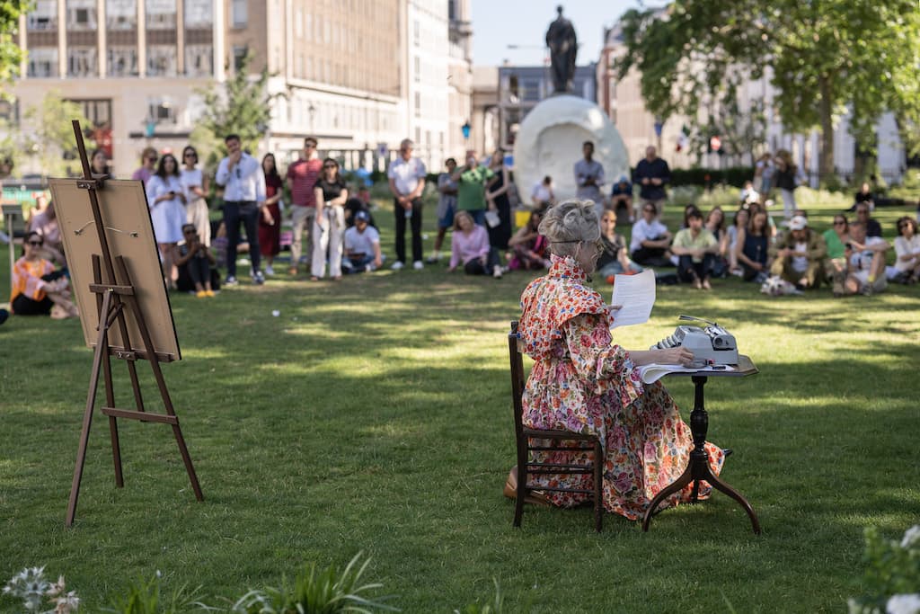 A performer in a floral dress sat at a wooden chair in the middle of a park reading some papers, with an audience watching the performance