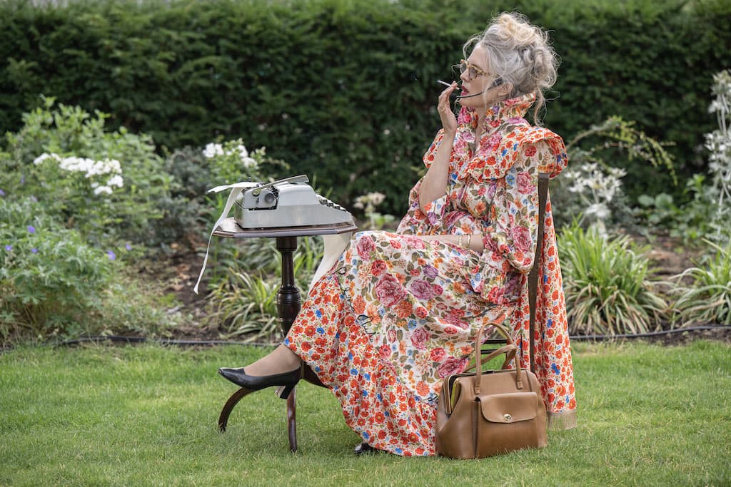A performer in a floral dress sat at a wooden chair and desk in the middle of a park smoking a cigarette.