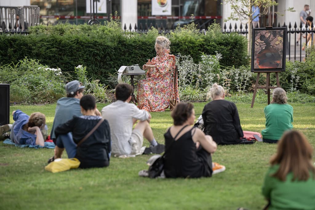 A performer in a floral dress sat at a wooden chair and desk in the middle of a park smoking a cigarette, with an audience in the foreground watching the performance.