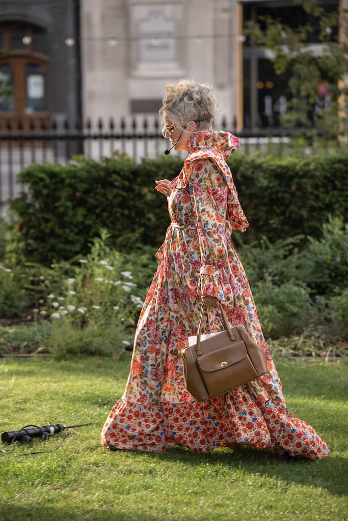 A performer in a floral dress with a microphone headset standing in middle of a park.