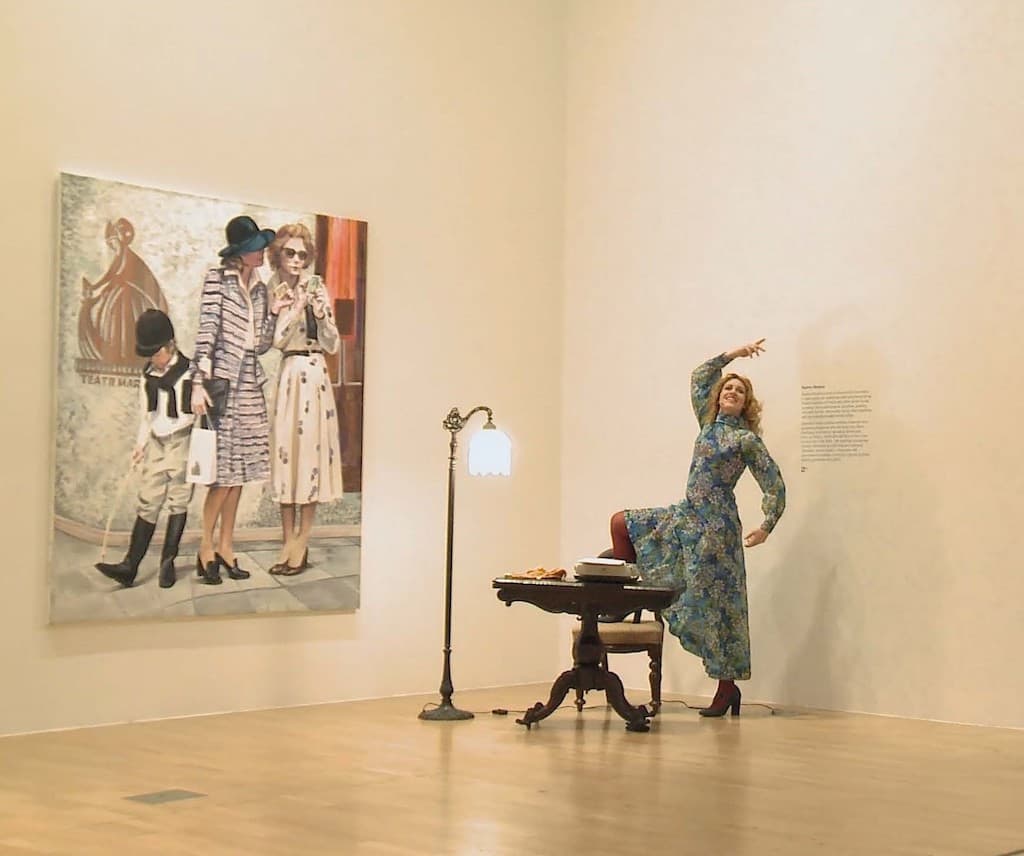 A performer in a floral dress pulling an elaborate pose within a small stage set installed in a gallery space with a large painitng on the wall.