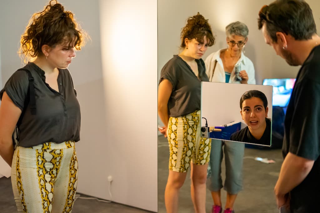 A performer sat in a mirrored booth, with audience in the foreground peering in.