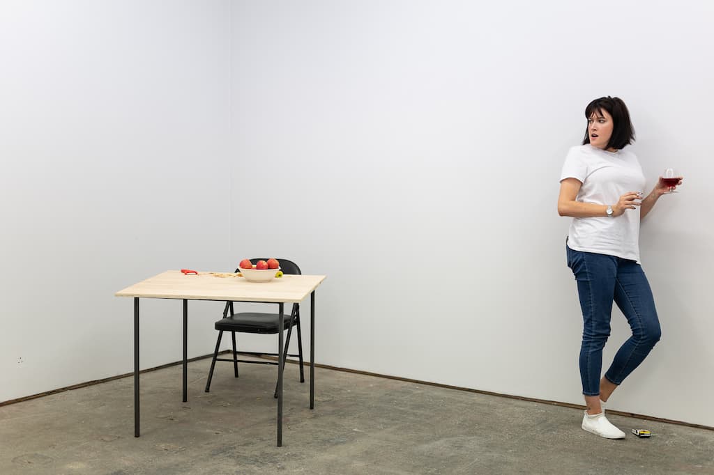 A performer leaning against a wall with a glass of red wine on one hand and a cigarette in the other. A table with a bowl of apples is in the background.