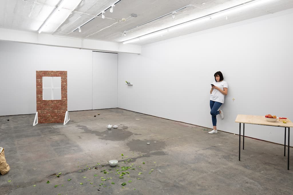 A performer leaning against a wall, texting. In the middle of the room is an empty white bowl with salad leaves scattered around it.