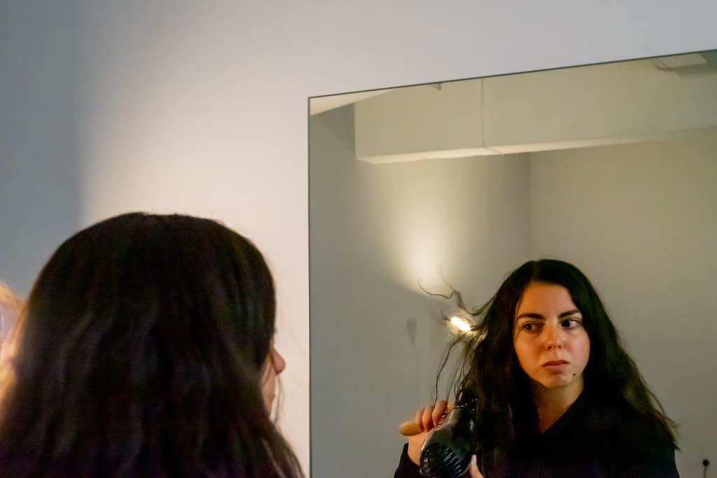 A performer looking into a mirror drying their hair with a hairdryer.