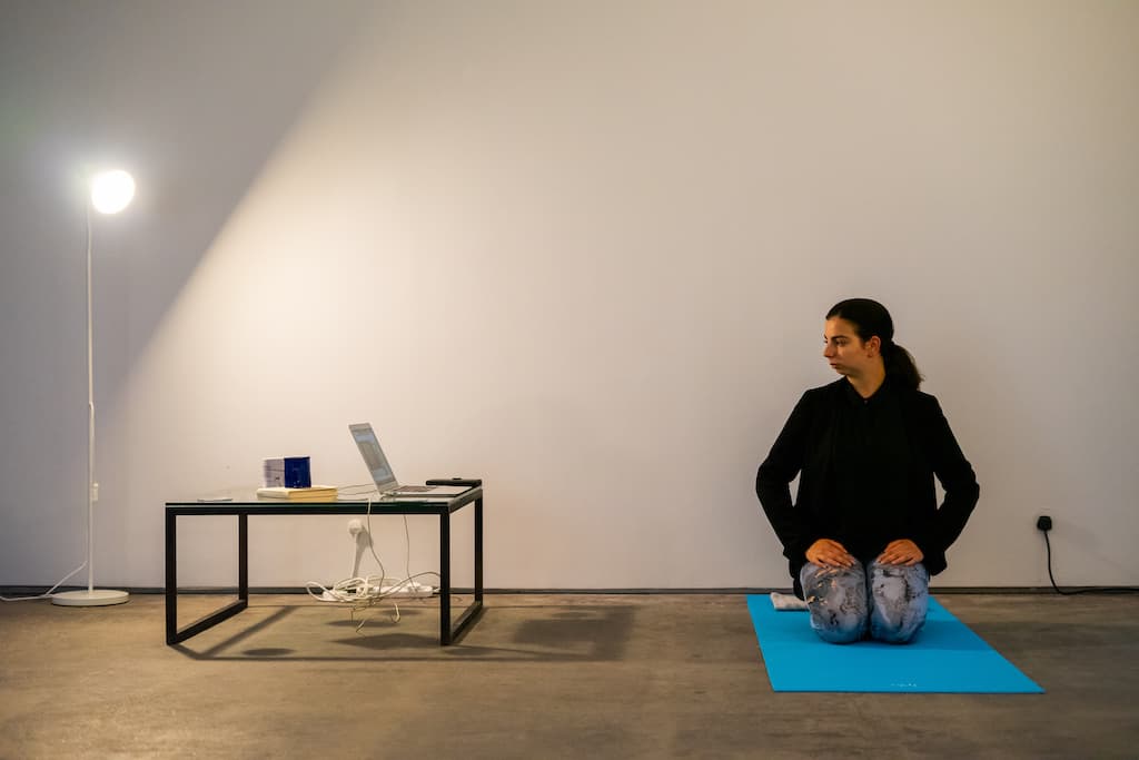 A performer kneeling on a yoga mat, looking to their right at a laptop on a low table