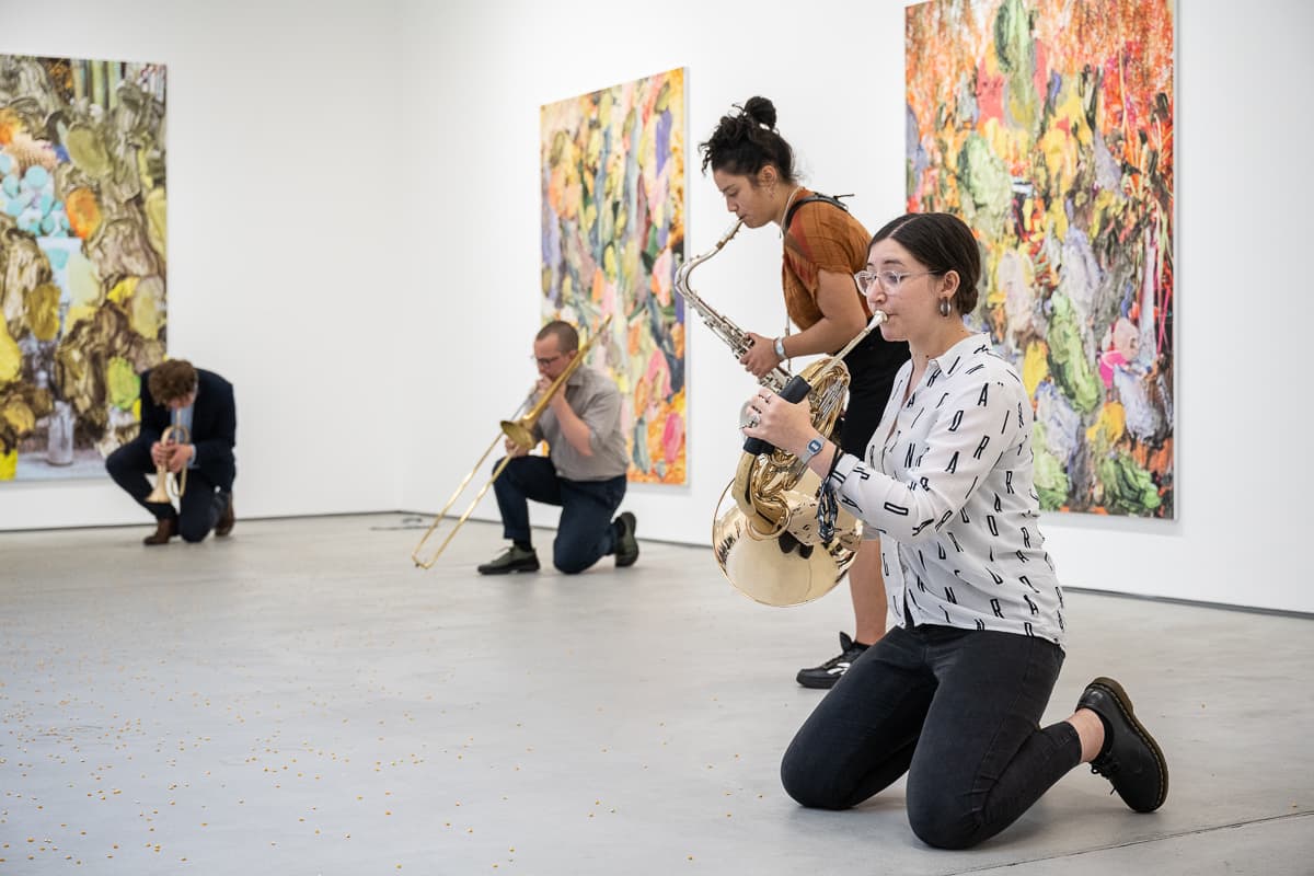 A group of musicans in a gallery, some sat on the floor, playing various instruments to un-popped popcorn kernals