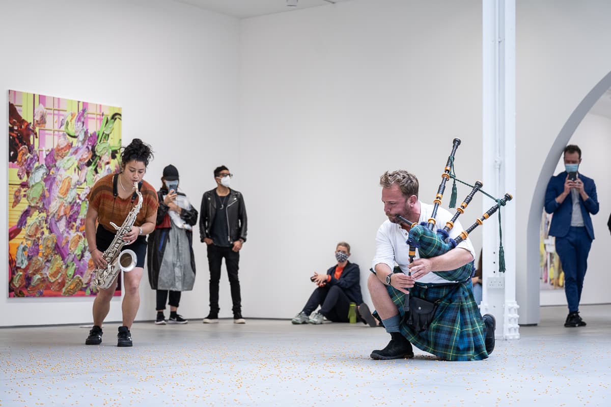 A group of musicans in a gallery, some sat on the floor, playing various instruments to un-popped popcorn kernals