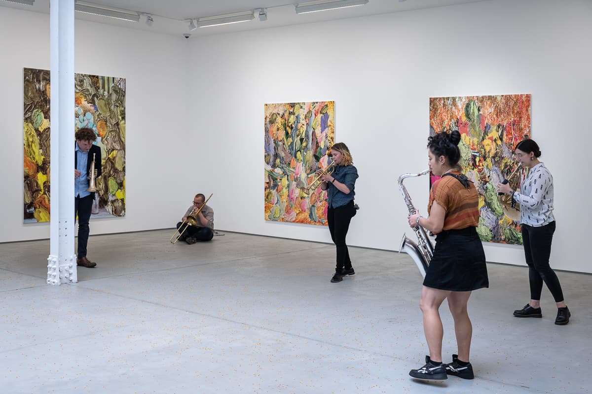 A group of musicans in a gallery, some sat on the floor, playing various instruments to un-popped popcorn kernals