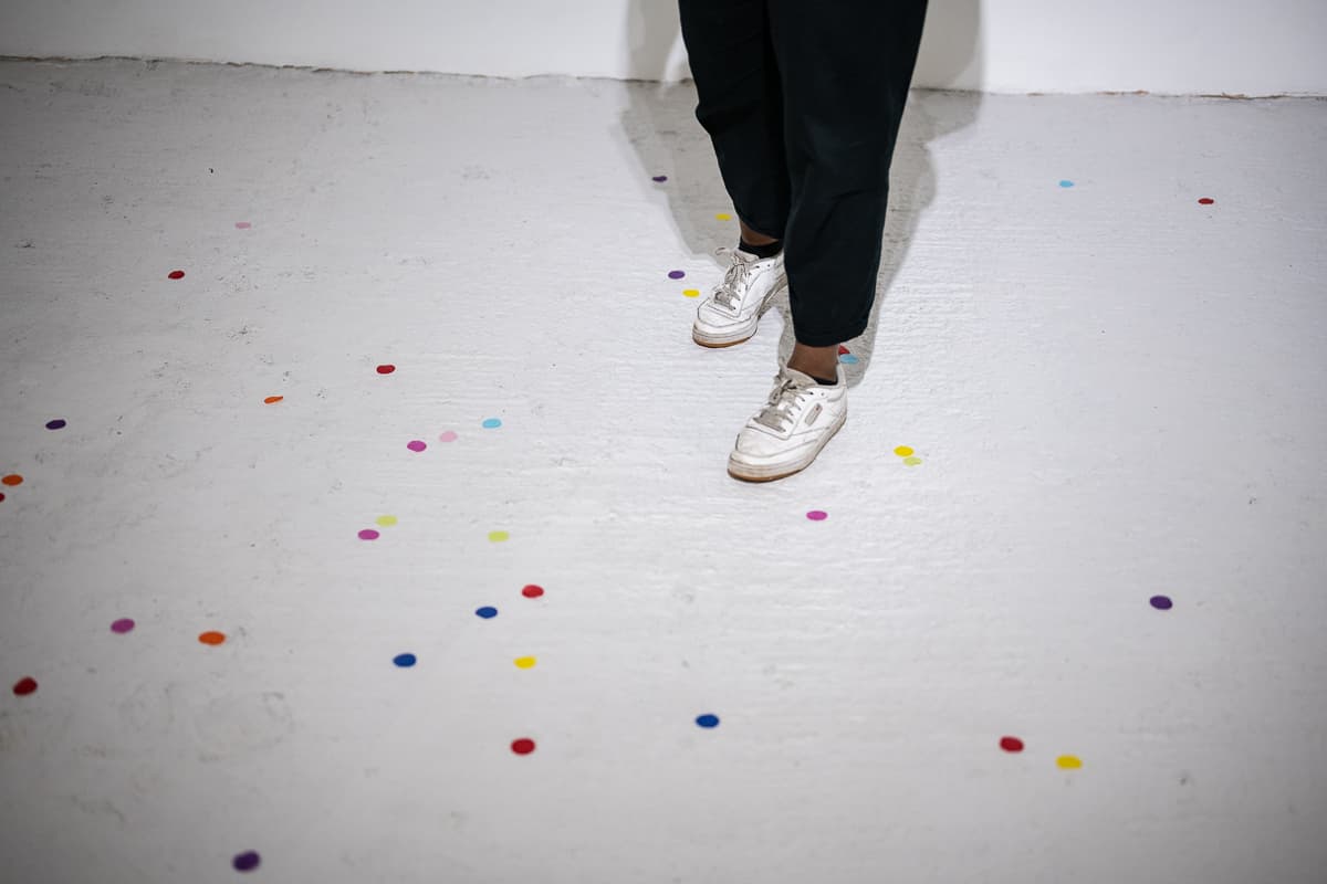 A female performer standing on the middle of a gallery space throwing peices of circular confetti to the ground.