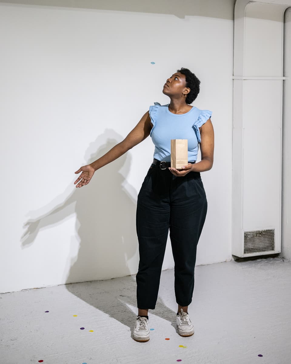 A female performer standing on the middle of a gallery space throwing peices of circular confetti to the ground.