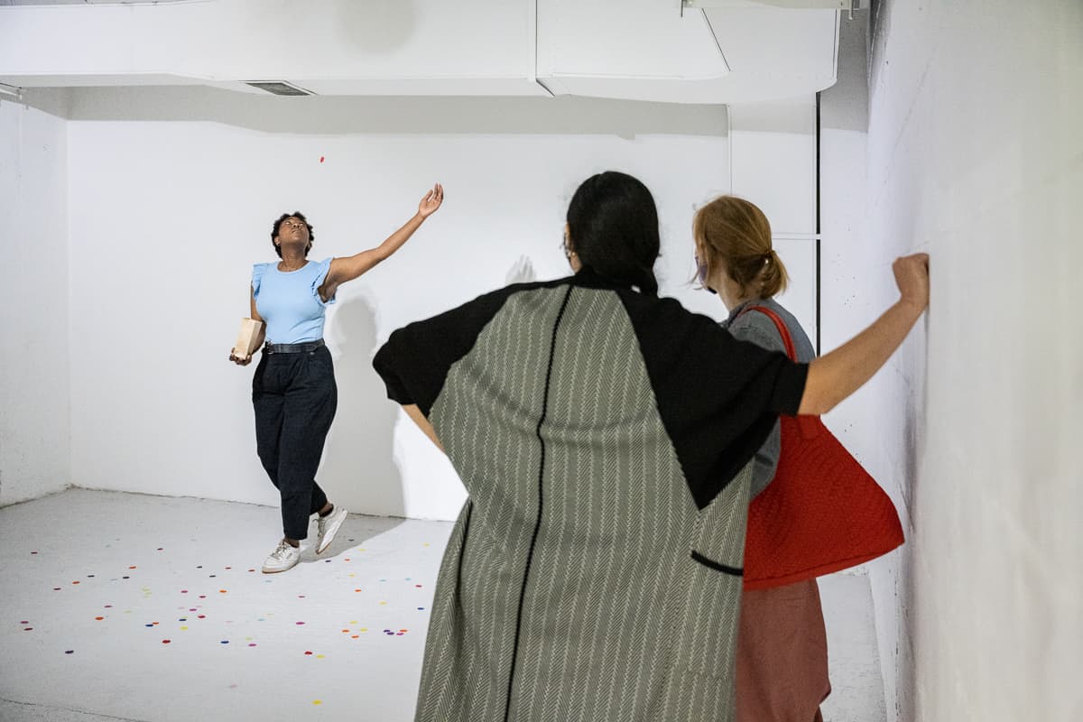 A female performer standing on the middle of a gallery space throwing peices of circular confetti to the ground.