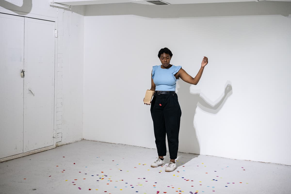 A female performer standing on the middle of a gallery space throwing peices of circular confetti to the ground.