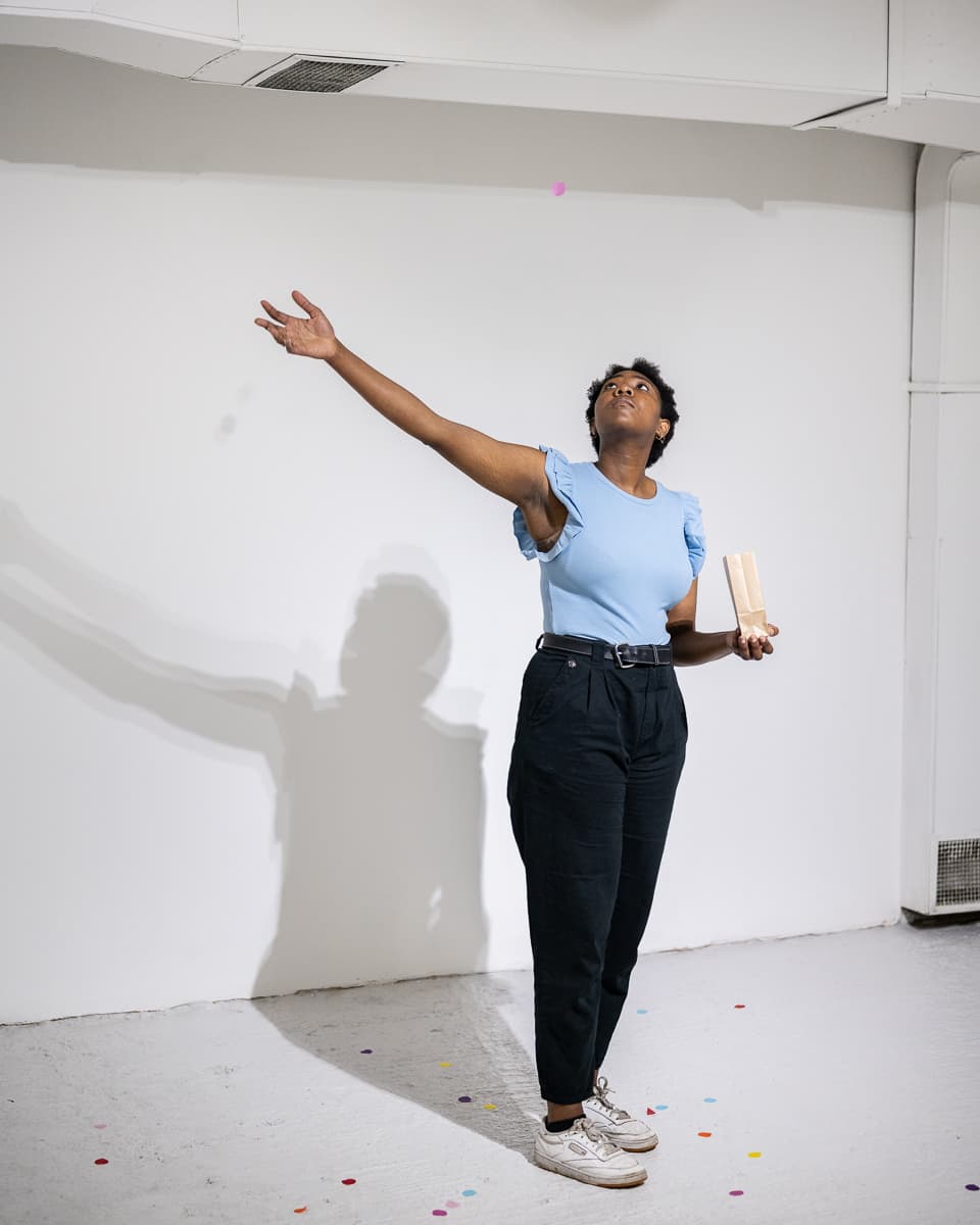 A female performer standing on the middle of a gallery space throwing peices of circular confetti to the ground.